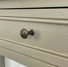 Load image into Gallery viewer, A close-up of a vintage-style round cabinet knob with a distressed dark metal finish, mounted on a light beige painted wooden drawer front with decorative molding.