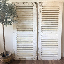 Load image into Gallery viewer, Pair of French vintage white wooden, chippy paint shutters on a wooden floor with a plant in the background.