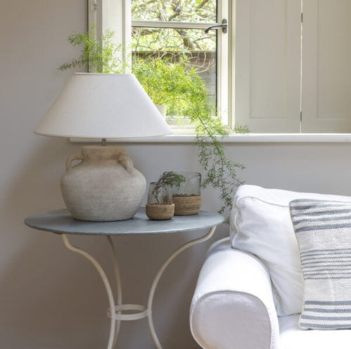 Lamp on a small table next to a white sofa with striped cushions, near a window with plants.