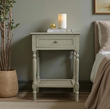 Load image into Gallery viewer, A light taupe nightstand with a single drawer and turned legs, styled in a bedroom next to a bed and a potted plant.